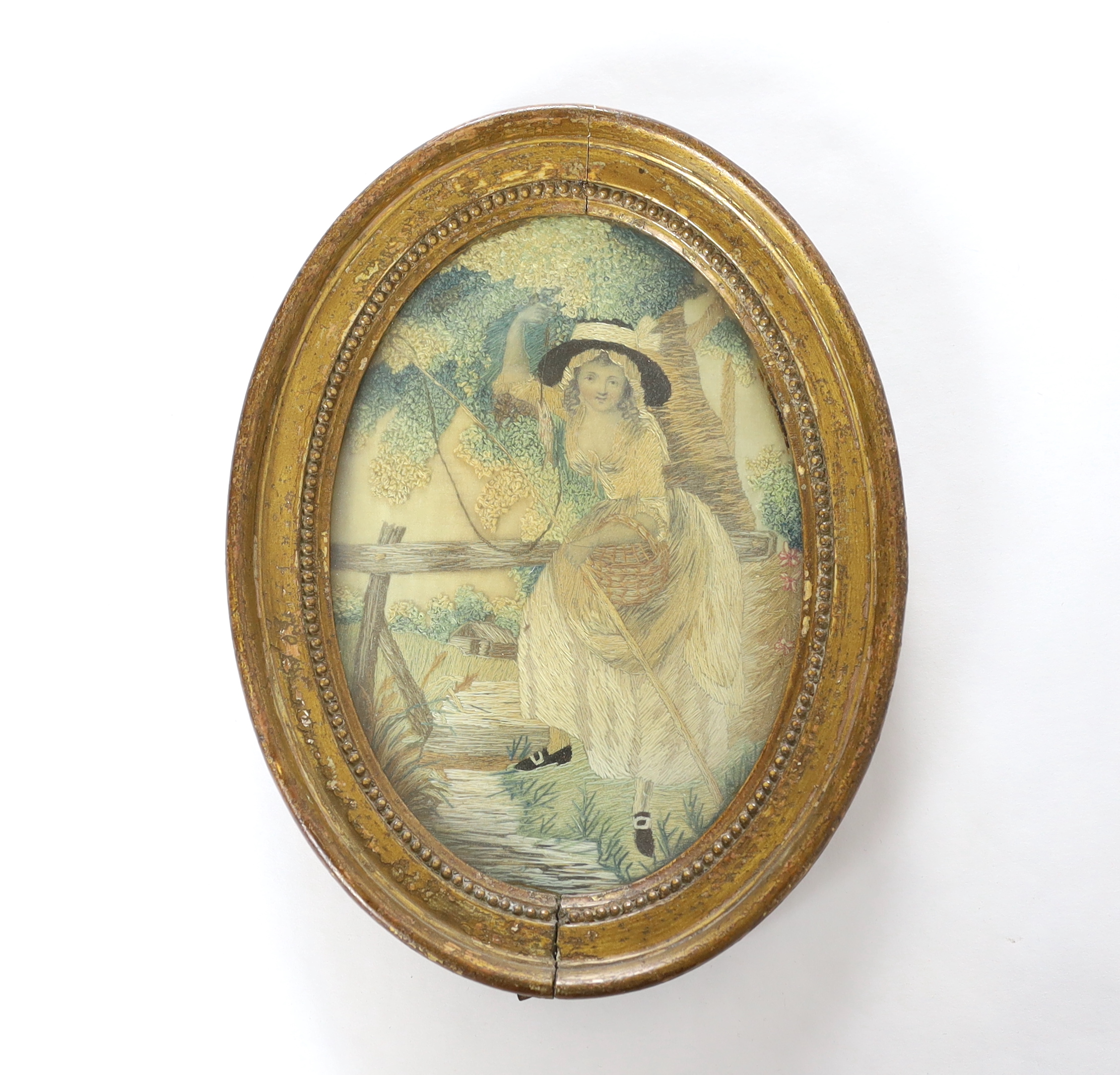 An early George IV oval silk work needlework pastoral embroidery, of a young girl with a fishing rod and basket under a tree by a bridge and stream, worked in fine wools and silks in stem stitch and a large knotting stit